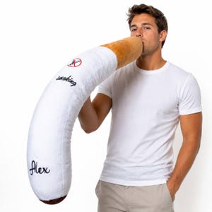 May include: A novelty cigarette-shaped pillow in white and beige. The pillow features the word "smoking" embroidered in black, along with a no-smoking symbol. The name "Alex" is also embroidered on the pillow.