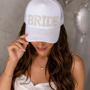 May include: A white trucker-style baseball cap featuring the word BRIDE in large, pearl-embellished block letters across the front panel. The cap has a solid white front and a mesh back, with a curved brim and a button at the top center.