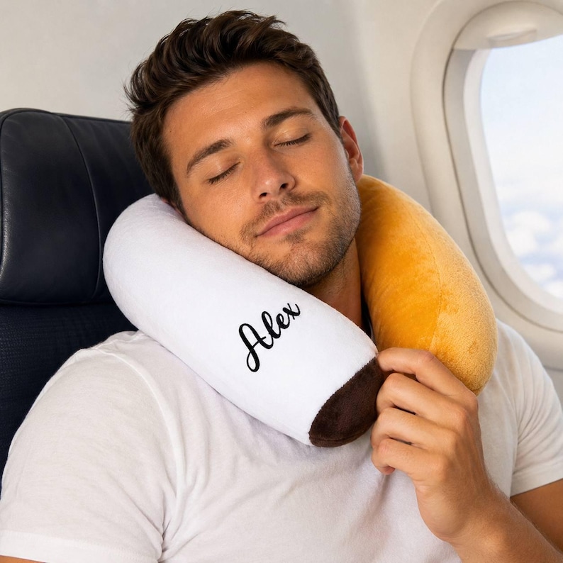 May include: A white and brown travel neck pillow with the name "Alex" printed on it. The pillow is around a person's neck, with a yellow pillow behind the neck. The person is wearing a white t-shirt and is seated on an airplane.