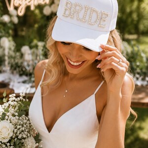 A smiling woman in a white dress and a white hat with the word "BRIDE" on it, holding a bouquet of white flowers.