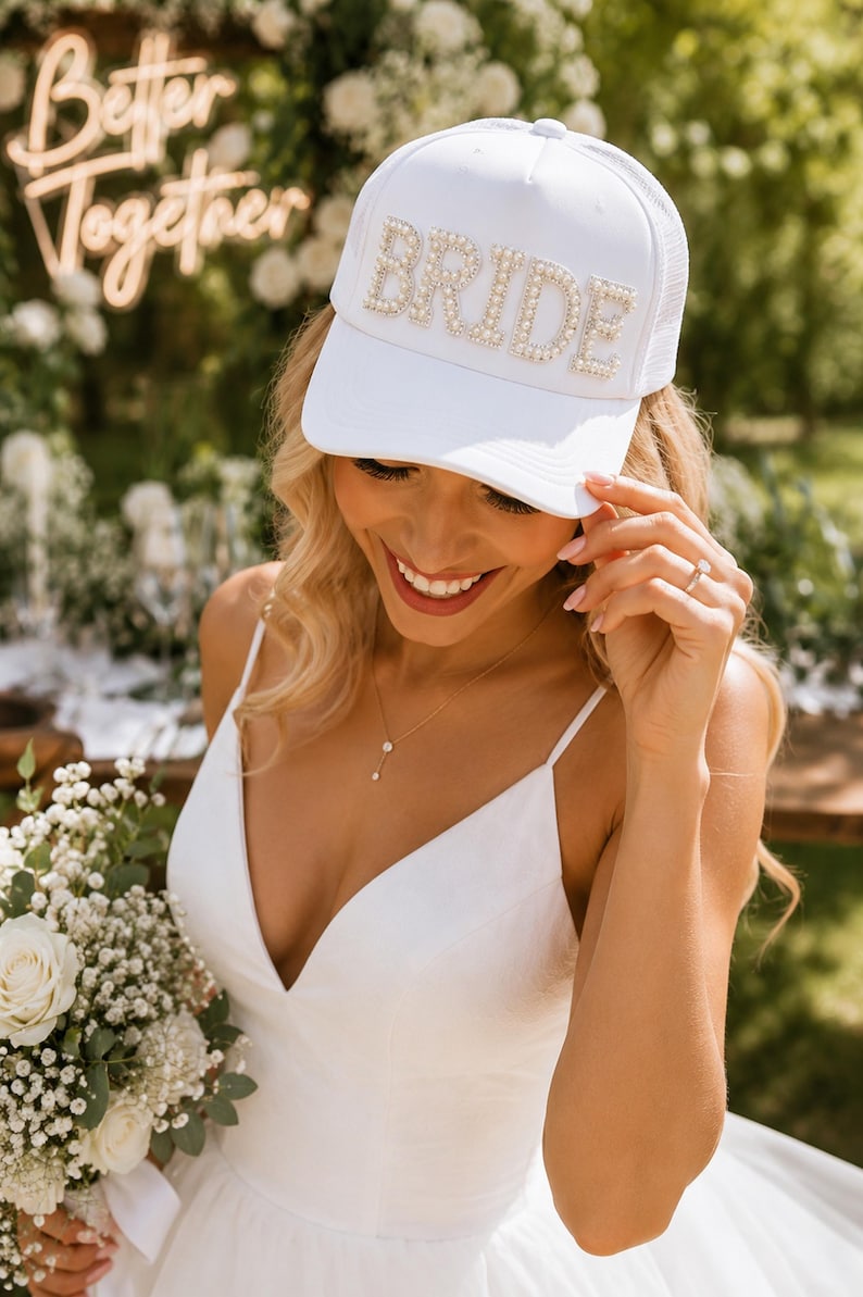 A smiling woman in a white dress and a white hat with the word "BRIDE" on it, holding a bouquet of white flowers.