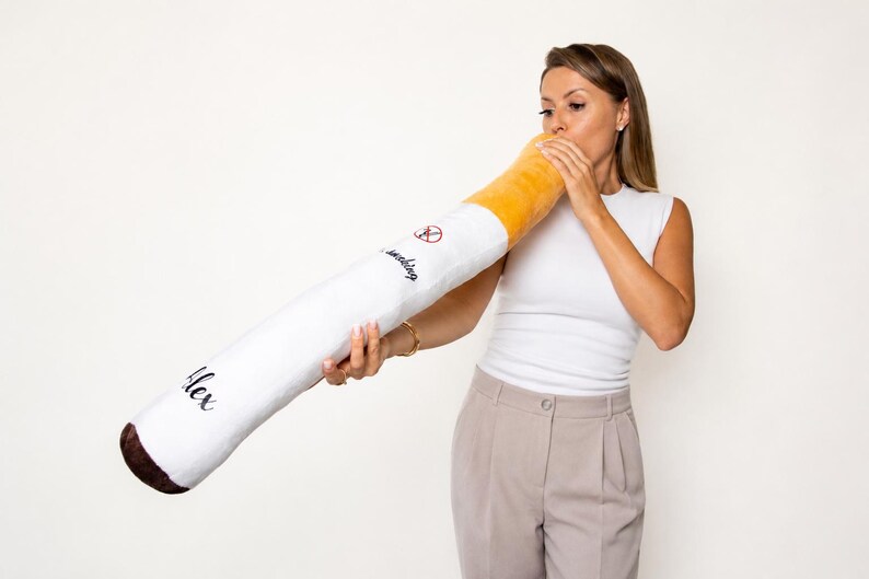 May include: A giant, soft cigarette-shaped cushion. It features a white body, a yellow tip, and a brown end. The cushion has the word "smoking" and a no-smoking symbol printed on it, along with the name "Alex".