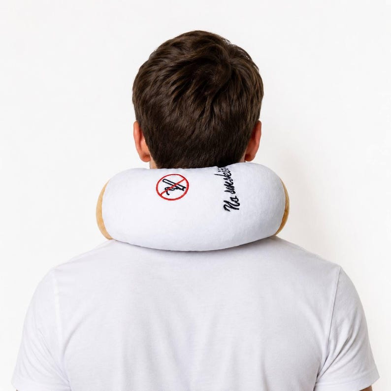 May include: A white travel neck pillow shaped like a cigarette, with tan ends. The pillow features a red "no smoking" symbol and the words "No smoking" embroidered in black. The person is wearing a white t-shirt.