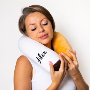 May include: A white and brown neck pillow with the name "Alex" in black script. The pillow has a yellow section and is being held by a person.