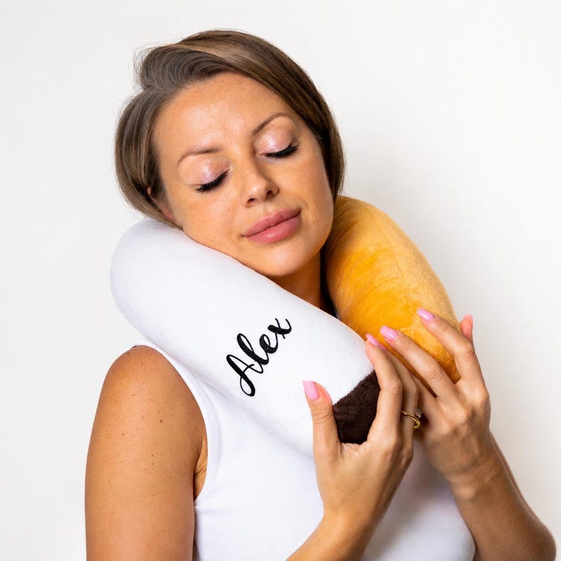 May include: A white and brown neck pillow with the name "Alex" in black script. The pillow has a yellow section and is being held by a person.