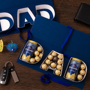 May include: A navy blue gift box with the word "DAD" in white, containing Ferrero Rocher chocolates and Sanpellegrino Limonata cans. Includes a black wallet, car keys, and cocktail umbrellas.