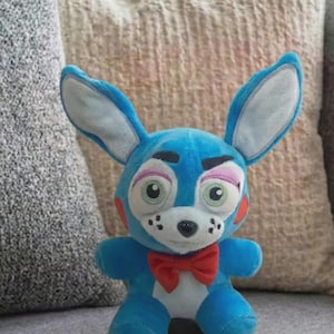 Puede incluir: Un peluche azul con forma de conejo con orejas grandes, una pajarita roja y un hocico blanco. El conejo tiene mejillas rosas, ojos verdes y blancos, y está sentado sobre una superficie gris.