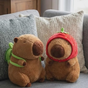 Könnte beinhalten: Zwei Plüsch-Wasserschweine sitzen auf einem grauen Sofa. Eines trägt einen grünen Rucksack, das andere einen roten Erdbeerhut. Beide haben braunes Fell, braune Nasen und geschlossene Augen. Der Hintergrund umfasst graue und cremefarbene Kissen.