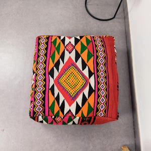 May include: A square floor cushion featuring a vibrant geometric pattern with diamonds and triangles in shades of orange, green, pink, black, and white. The textile surface displays a repeating diamond motif with a central yellow and orange grid.