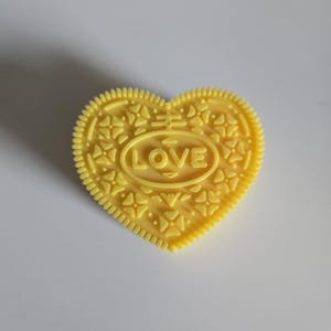 May include: A yellow heart-shaped biscuit with the word "LOVE" embossed in the centre. The biscuit has a detailed pattern around the edges and on the surface. The biscuit is a bright yellow colour.