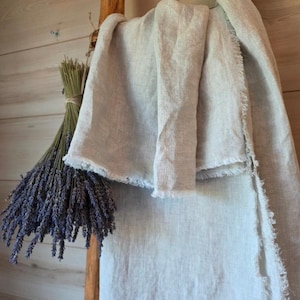 May include: A light beige linen throw blanket featuring a textured weave and frayed edges, displayed alongside a tied bundle of dried purple lavender stems. The fabric has a natural, slightly wrinkled appearance with visible fibers and loose threads.
