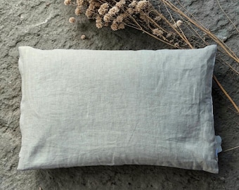 May include: A rectangular throw pillow with a light beige, textured fabric cover. The pillow has a visible seam along the edge and a small white tag on the side. The surface of the fabric shows a woven, natural appearance with subtle color variations.