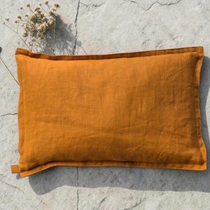 May include: Mustard-colored rectangular pillow made of linen fabric. The pillow has a simple design with a stitched border and a small tab on one side. The pillow is approximately 18 inches long and 12 inches wide.