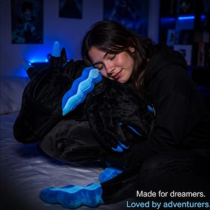 Op de afbeelding: Een pluche draak met zwarte vacht en felblauwe accenten op de hoorns, manen en klauwen. De tekst op de afbeelding luidt: Made for dreamers. Loved by adventurers.
