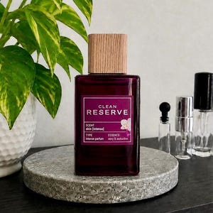 May include: A rectangular glass perfume bottle with a deep purple tint and a light-colored wooden cap. The label reads CLEAN RESERVE, SCENT skin [intense], TYPE ESSENCE, intense parfum, and sexy & seductive, featuring a small white orchid graphic.