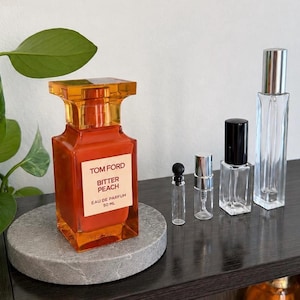 May include: A perfume bottle with a rectangular shape and an orange hue, labeled "Bitter Peach" by Tom Ford, sits on a gray stone. Several clear glass perfume sample bottles of varying sizes are also present, with silver and black caps.