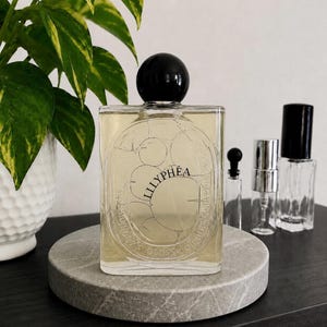 May include: A rectangular glass perfume bottle with a black spherical cap, containing a light yellow liquid. The front label features the text LILLYPHÉA in black lettering, surrounded by a circular line drawing of lily pads and text.