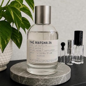 May include: A clear glass perfume bottle with a silver cap and a white label reading THÉ MATCHA 26, 100ml 3.4 FL.OZ. e, eau de parfum, natural spray, labelled in SSG Main DF byMA on 08/15/26 for You, with three smaller empty glass vials nearby.