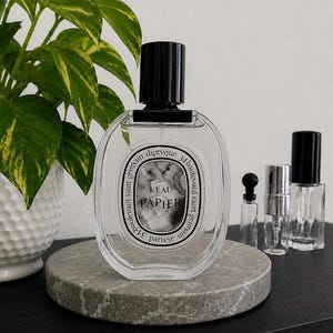 May include: A clear glass perfume bottle with a black cap and a white oval label featuring the text L'EAU PAPIER and diptyque 34 boulevard saint germain. Three smaller empty glass vials with black and silver caps are positioned to the right side.
