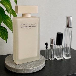 May include: A large rectangular frosted glass perfume bottle with a beige cap, labeled PURE MUSC BLANC narciso rodriguez for her, accompanied by four smaller clear glass spray bottles of varying heights with black and silver caps.