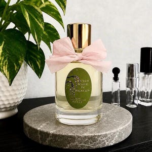 May include: A clear glass perfume bottle with a gold cap and a light pink fabric bow tied around the neck. The bottle features an oval green label with the text Match Made In Heaven and 50 ml printed on it. The liquid inside is pale yellow.