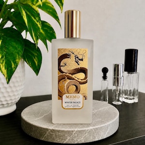 May include: A rectangular frosted glass perfume bottle with a gold cap. The front label features a gold background with a black dragon illustration and the text MEMO PARIS WINTER PALACE EAU DE PARFUM graines vagabondes in black lettering.