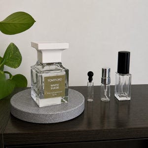 May include: A clear glass perfume bottle with a white cap and a label that reads "TOM FORD WHITE SUEDE EAU DE PARFUM 30 ML" sits on a gray stone coaster. Several smaller perfume sample bottles are also present.