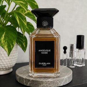 May include: A rectangular glass perfume bottle filled with amber liquid, featuring a black label with the white text ANGÉLIQUE NOIRE and GUERLAIN PARIS. The bottle has a faceted design and a black cap with a textured neck and a circular logo.