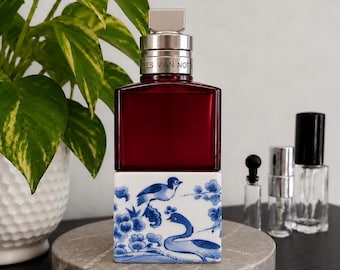 May include: A Dries Van Noten perfume bottle with a deep red glass upper section and a white ceramic base featuring blue painted illustrations of birds and floral branches. The silver metallic cap is engraved with the brand name.