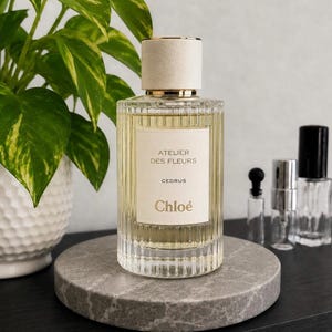 May include: A clear glass perfume bottle with vertical fluting and a cream-colored cap. The label features the text ATELIER DES FLEURS, CEDRUS, and Chloé. The bottle contains a light yellow liquid and is positioned on a circular gray stone base.