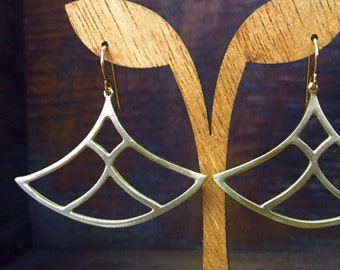 May include: A pair of gold-toned earrings with a geometric fan-shaped design. The earrings are hanging from a wooden display stand.