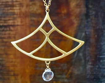 May include: A gold necklace with a geometric pendant and a clear crystal drop. The pendant is shaped like a fan and has a delicate, intricate design.