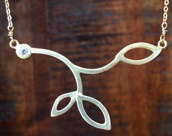 May include: A gold necklace with a delicate branch design and a single clear gemstone. The branch has three leaves on one side and one leaf on the other.