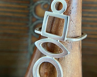 May include: A silver ring with a geometric design featuring a circle, square, and oval shapes. The ring is displayed on a wooden stand.