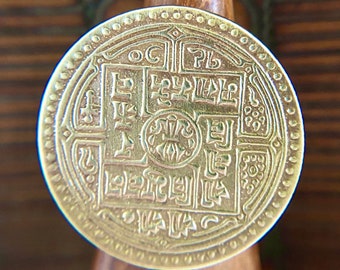 May include: A gold-colored metal ring with intricate designs and lettering. The ring is circular and has a raised edge. The design features a central square with four smaller squares surrounding it. Each square contains lettering and decorative elements. The ring is sitting on a wooden surface.