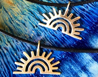 May include: Two gold-toned necklaces with a sun and rainbow design pendant. The necklaces are on a black cord and are displayed on a blue and gold background.
