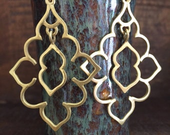 May include: A pair of gold-tone earrings with a geometric design. The earrings are hanging from a ceramic vase with a brown and green glaze.