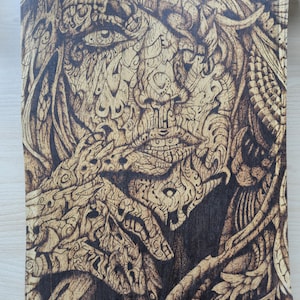 May include: A detailed wood-burned artwork depicting a woman's face with intricate patterns. The artwork features a light brown color with darker brown shading, creating depth and highlighting the facial features and flowing hair. The woman's hand is near her face.