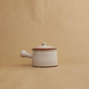 May include: A small, off-white ceramic pot with a single side handle and a matching lid. The pot features a distinct reddish-brown rim around the top edge and a matching band at the base, providing a subtle contrast to the light-colored body.