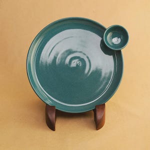 Peut inclure: Une assiette ronde en céramique de couleur sarcelle avec une finition lisse et brillante, dotée d'un petit bol circulaire assorti fixé sur le bord supérieur droit. L'assiette est maintenue par un support en bois brun foncé.