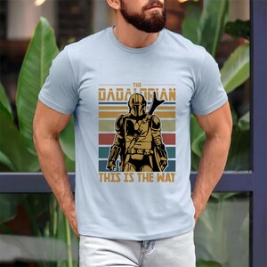 May include: Pale blue t-shirt with a retro graphic of a Mandalorian warrior. The design includes the text "THE DADALORIAN" and "THIS IS THE WAY", along with a helmeted figure and horizontal stripes in various colours.