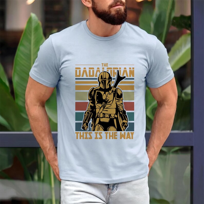 May include: Pale blue t-shirt with a retro graphic of a Mandalorian warrior. The design includes the text "THE DADALORIAN" and "THIS IS THE WAY", along with a helmeted figure and horizontal stripes in various colours.