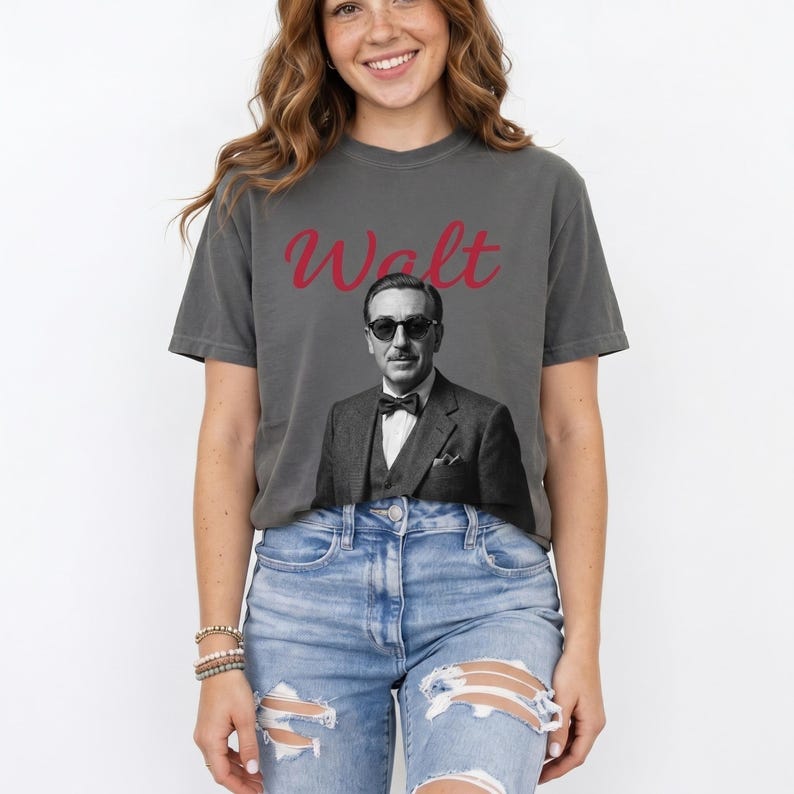 A front view photograph of a smiling young woman with wavy brown hair, wearing a charcoal grey graphic t-shirt. The shirt features a large, black and white portrait photograph of a man resembling Walt Disney in a suit and sunglasses, with "Walt" written above it in red cursive text. She is also wearing distressed, light-wash denim jeans and stacked beaded bracelets on her wrist, standing against a plain white studio background.