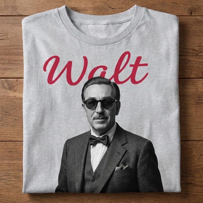Light gray t-shirt laid flat on a wooden surface, featuring a retro black and white portrait of Walt Disney in sunglasses with the word "Walt" in red cursive text. A perfect retro Walt Disney shirt for fans.