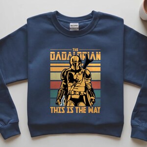 May include: Navy blue sweatshirt with a graphic of a Mandalorian-style character. The text reads "THE DADALORIAN" and "THIS IS THE WAY" in yellow. The design includes retro stripes in orange, green, and red.