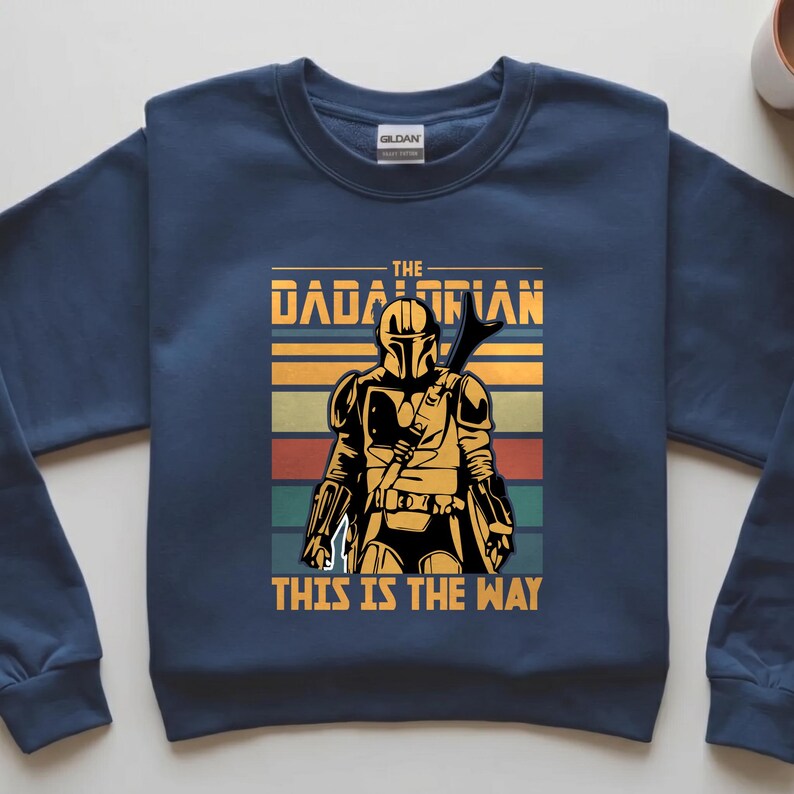 May include: Navy blue sweatshirt with a graphic of a Mandalorian-style character. The text reads "THE DADALORIAN" and "THIS IS THE WAY" in yellow. The design includes retro stripes in orange, green, and red.