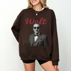 A female model wearing a dark brown oversized Happiest Place On Earth sweatshirt, featuring a classic black and white portrait of Walt Disney wearing sunglasses with red signature-style text.