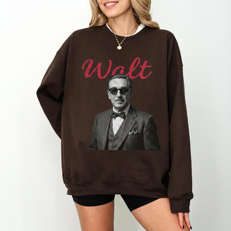 A female model wearing a dark brown oversized Happiest Place On Earth sweatshirt, featuring a classic black and white portrait of Walt Disney wearing sunglasses with red signature-style text.