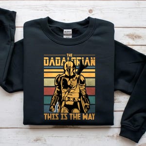 May include: Black sweatshirt with a graphic of a Mandalorian character. The text reads "THE DADALORIAN" and "THIS IS THE WAY". The design uses a retro colour palette of gold, orange, green, and blue.