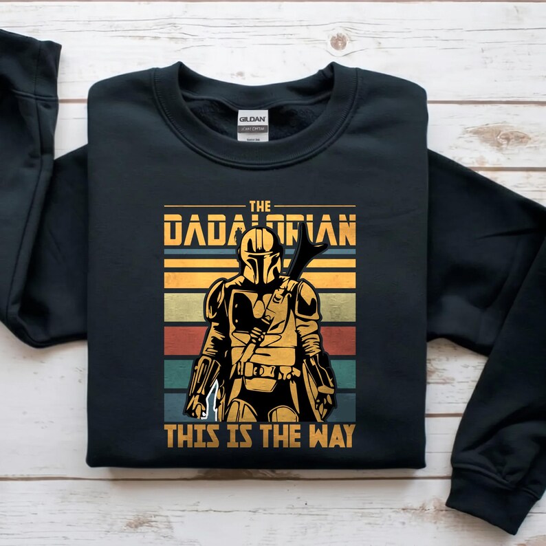 May include: Black sweatshirt with a graphic of a Mandalorian character. The text reads "THE DADALORIAN" and "THIS IS THE WAY". The design uses a retro colour palette of gold, orange, green, and blue.